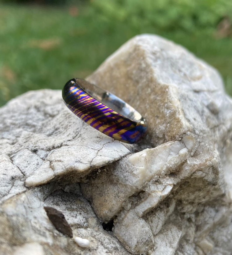 May include: A close-up of a colourful, patterned ring with a silver band. The ring is made of a metal with a unique, iridescent finish. The ring is resting on a grey rock.