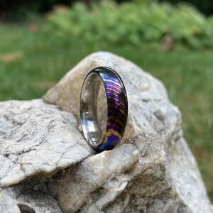 Op de afbeelding: Een zilveren en iriserende ring met een regenboogpatroon. De ring ligt op een grijze steen.