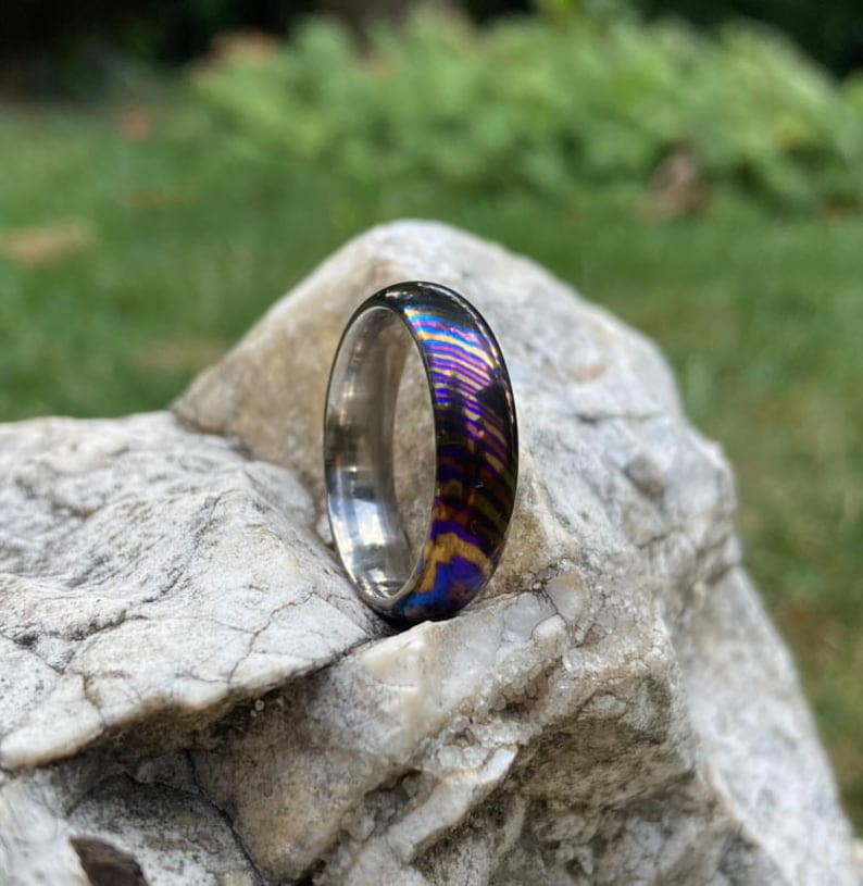 May include: A silver and iridescent ring with a rainbow pattern. The ring is sitting on a grey rock.
