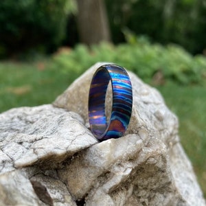 May include: A blue and purple patterned metal ring, sitting on a gray rock.