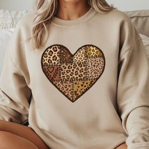 Puede incluir: Sudadera beige con un gran diseño de corazón. El corazón está compuesto por varios estampados de leopardo y patchwork en tonos marrones y dorados. La sudadera es de estilo casual y cómodo.