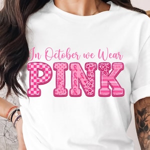 Könnte beinhalten: Ein weißes T-Shirt mit kurzen Ärmeln und dem rosa Schriftzug In October we Wear PINK. Das Wort PINK besteht aus großen Blockbuchstaben, die jeweils mit verschiedenen Mustern wie Karos, Schleifen, Herzen und Leopardenmuster versehen sind.