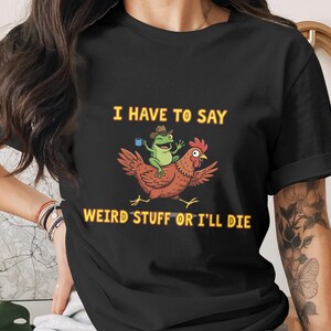Puede incluir: Camiseta negra con un gráfico humorístico. El diseño presenta una gallina de dibujos animados con una rana montada en su espalda, sosteniendo una taza de café. El texto dice "I HAVE TO SAY WEIRD STUFF OR I'LL DIE" en amarillo.