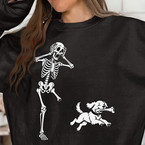 May include: A black long-sleeve sweatshirt featuring a white graphic print of a skeleton standing with hands on its head in a shocked pose, positioned next to a small dog running while carrying a large bone in its mouth.