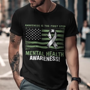 Puede incluir: Camiseta negra con un diseño gráfico verde y blanco. El diseño incluye una bandera estadounidense, una cinta y el texto "AWARENESS IS THE FIRST STEP" y "MENTAL HEALTH AWARENESS!"