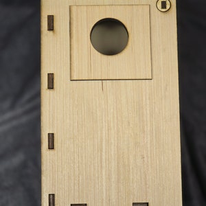 Pode incluir: Uma casa de pássaros de madeira com uma abertura redonda e uma pequena janela retangular. A casa tem um telhado marrom e é feita de madeira clara.