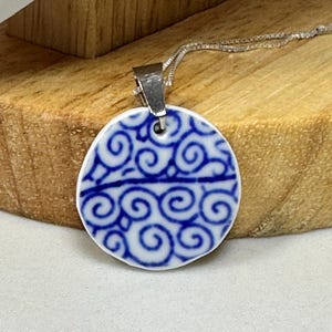 May include: A round, white and blue pendant necklace. The pendant features a blue swirl design on a white background. The necklace has a silver chain and clasp. The pendant is displayed on a wooden stand.