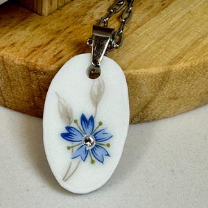 May include: Oval white pendant necklace with a silver chain. The pendant features a blue flower design with a small crystal in the center. The flower is surrounded by two gray leaves.