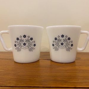 May include: Two white ceramic mugs with a navy blue floral design. Each mug has a square handle and a flared shape. The floral design is a stylized tree with flowers and leaves. The mugs are on a wooden surface.
