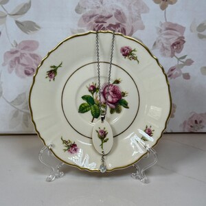 Op de afbeelding: Een crèmekleurige hangerketting met een zilveren ketting en een kleine, ovaalvormige hanger met een roze rozenontwerp. De ketting wordt getoond op een vintage bord met een gouden rand en rozenpatroon.