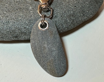 Könnte beinhalten: Ein grauer, glatter Stein-Schlüsselanhänger mit einem silberfarbenen Verschluss. Der Stein ist oval geformt und hat einen Metallring, der am Verschluss befestigt ist. Das Accessoire ist natürlich und minimalistisch.