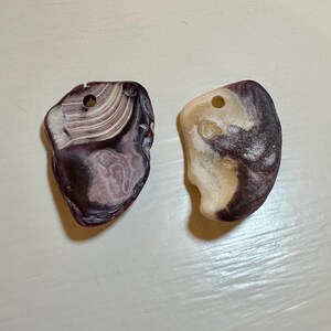 May include: Two purple and white seashells with holes drilled through them. The shells are smooth and have a natural, organic shape.