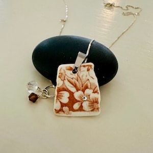 May include: A brown and white porcelain pendant with a floral design and a single rhinestone. The pendant is attached to a silver chain.