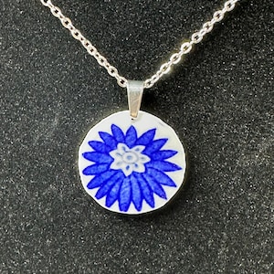 May include: A silver chain necklace with a round, white pendant. The pendant features a blue floral design in the centre. The flower has a detailed pattern with blue petals and a white centre. The necklace is displayed against a dark background.