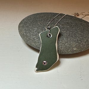 May include: A green ceramic pendant with a pink rhinestone and a silver chain. The pendant is shaped like a foot and has a white border.