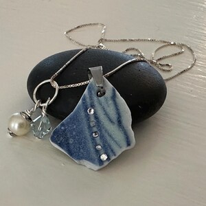 May include: A silver chain necklace with a blue and white ceramic pendant featuring a swirl design and clear rhinestones. The pendant is attached to a silver ring with a pearl and a blue crystal bead.