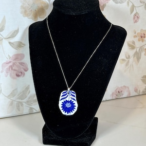May include: A blue and white floral pendant necklace on a silver chain. The pendant is a unique shape with a blue floral design. The necklace is displayed on a black jewelry bust against a floral background.
