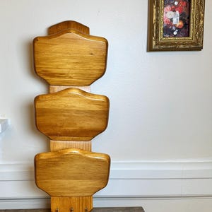 May include: A vertical wooden wall organizer featuring three stacked, rounded rectangular shelves with a smooth, light brown finish. The structure is mounted on a central wooden spine, and three small metal hooks are attached to the bottom section.