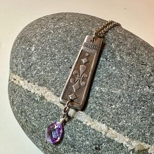 May include: A silver pendant necklace with a rectangular pendant featuring an embossed design and a teardrop-shaped purple gemstone. The pendant hangs from a delicate chain. The necklace is displayed on a gray stone.