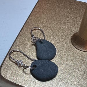 May include: A pair of handcrafted earrings featuring dark gray, oval-shaped stones. Each earring has a silver-colored hook and a small, clear crystal bead. The earrings are displayed on a gold-colored surface.