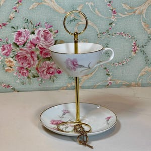 May include: A two-tiered serving stand with a gold-colored metal frame. The top tier is a teacup, and the bottom tier is a saucer, both decorated with pink floral patterns. The stand is designed for displaying small items.