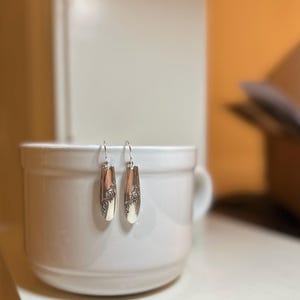 Può includere: Un paio di orecchini d'argento con un design floreale. Gli orecchini sono appesi a una tazza di ceramica bianca.
