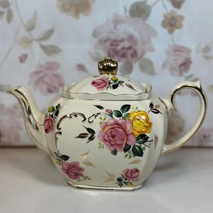 Könnte beinhalten: Cremefarbene Vintage-Teekanne mit einem floralen Rosendesign in Rosa und Gelb. Die Teekanne hat goldfarbene Verzierungen und einen goldfarbenen kronenförmigen Knauf auf dem Deckel. Griff und Ausguss sind ebenfalls goldfarben.