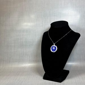 May include: A silver necklace with a round pendant featuring a blue and white floral design. The pendant is displayed on a black velvet jewellery bust against a neutral background.