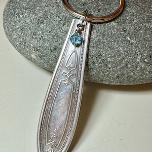 Puede incluir: Un llavero de cuchara de plata con un dije de cristal azul. La cuchara tiene un diseño adornado y está unida a un llavero de plata.
