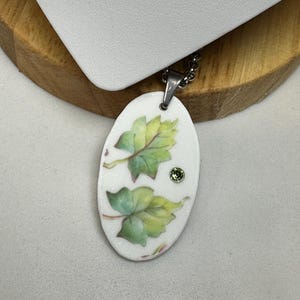 May include: Oval pendant necklace with a white background, featuring green and yellow leaf illustrations and a small green gem. The pendant hangs from a silver chain. The necklace is displayed on a wooden and white surface.