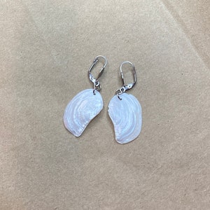 以下が含まれることがあります： 銀色のフックが付いた白い貝殻のイヤリングのペア。貝殻は自然な虹色の仕上げになっています。