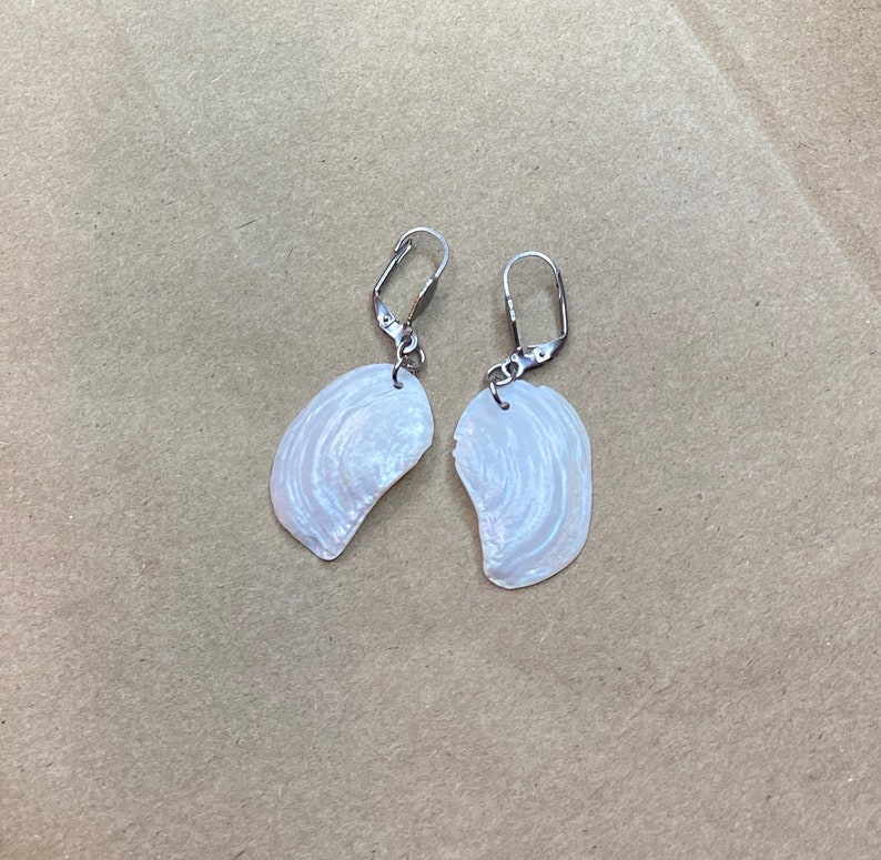以下が含まれることがあります： 銀色のフックが付いた白い貝殻のイヤリングのペア。貝殻は自然な虹色の仕上げになっています。