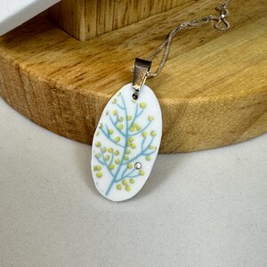 May include: Oval pendant necklace with a silver chain. The white pendant features a blue and yellow floral design with a small, clear gemstone. The necklace is displayed on a wooden stand.