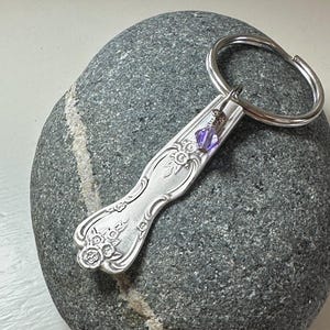 Puede incluir: Un llavero de plata hecho con una pieza de cubiertos vintage con un dije de cristal morado. El llavero está apoyado sobre una piedra gris.