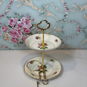 May include: Two-tiered, off-white ceramic serving dish with a gold-colored handle and stand. Each dish features a floral pattern with pink roses and green leaves. A gold bracelet and key chain rest on the bottom tier.