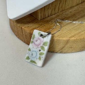 Puede incluir: Un collar con colgante rectangular con un diseño floral. El colgante presenta rosas rosas y azules con hojas verdes sobre un fondo blanco. Una pequeña gema transparente está engastada debajo de la rosa azul. El collar tiene una delicada cadena de plata.