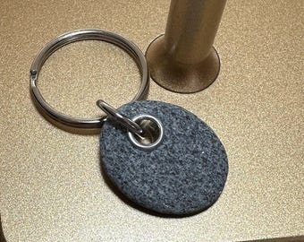 Könnte beinhalten: Ein grauer, oval geformter Schlüsselanhänger mit einem silberfarbenen Spaltring. Der Anhänger ist an einem größeren silbernen Schlüsselring befestigt. Der Hintergrund ist eine goldfarbene Oberfläche.