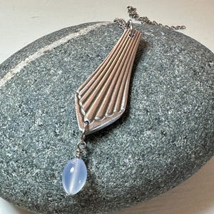 May include: A silver necklace with a long, thin, fan-shaped pendant and a light blue oval-shaped bead hanging from the bottom.