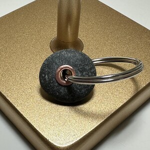 May include: A dark gray stone keychain with a copper-colored grommet and a silver-colored key ring. The keychain is resting on a gold-colored, textured surface.