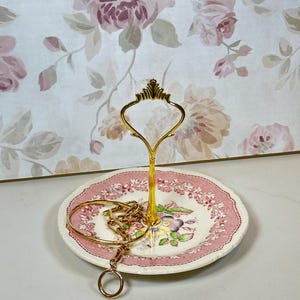 May include: A two-tiered jewelry stand with a gold-colored handle and a floral patterned plate. The plate has a pink border and a floral design. A gold bracelet and necklace rest on the plate. The background features a floral wallpaper.
