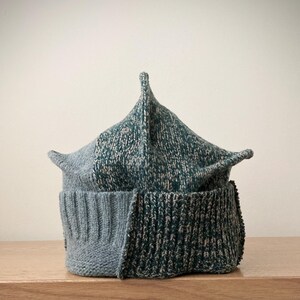 Puede incluir: Un gorro de punto con corona puntiaguda y borde doblado. El gorro está construido con dos secciones distintas: un lado es de color azul claro sólido, mientras que el otro lado es una mezcla moteada de hilo verde azulado oscuro y gris claro.
