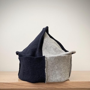 May include: A two-toned beanie hat constructed from dark navy blue and light heather gray fabric panels. The hat features a pointed crown design with visible stitched seams connecting the contrasting colored sections and a ribbed knit base.