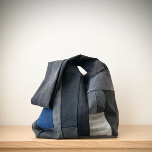 May include: A patchwork tote bag in shades of gray and blue. The bag features a variety of fabric textures and patterns, with a large handle and a rectangular shape. The bag is sitting on a wooden surface.