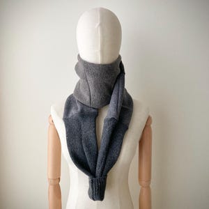 Puede incluir: Bufanda gris de dos tonos exhibida en un maniquí. La bufanda está hecha de un material suave y tejido. La parte superior es de un gris más claro, que pasa a un gris más oscuro en la mitad inferior. La bufanda está diseñada para usarse alrededor del cuello.