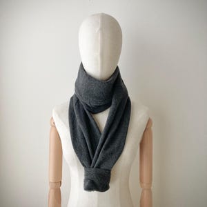 May include: A dark gray infinity scarf displayed on a mannequin. The scarf is made of a soft, knitted material and is draped around the mannequin's neck and shoulders. The scarf's design is simple and versatile, suitable for various styles.