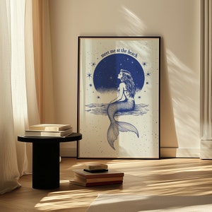 Peut inclure: Une impression encadrée avec une illustration bleue d'une sirène assise sur un rocher avec une pleine lune et des étoiles en arrière-plan. Le texte "Meet me at the beach" est écrit en bleu.