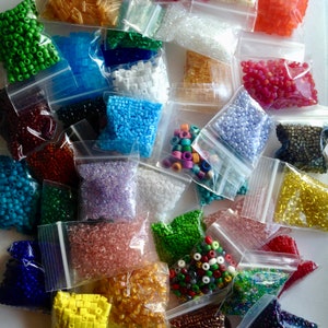 May include: A collection of numerous small, clear plastic resealable bags filled with assorted craft beads in a wide variety of colors, shapes, and finishes, including opaque, translucent, metallic, and iridescent styles scattered across the frame.