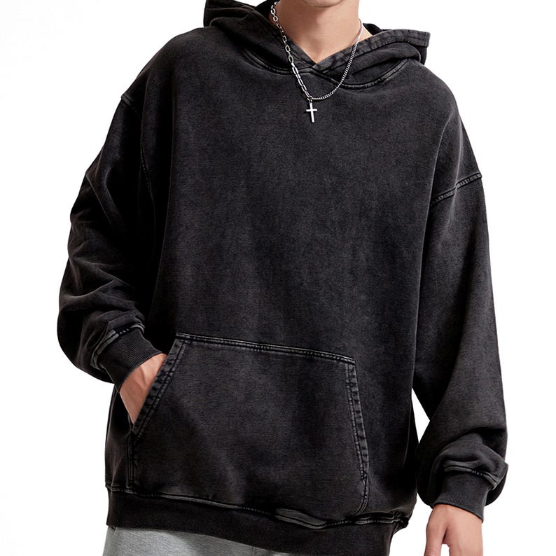 May include: A black, washed-out hoodie with a kangaroo pocket and a drawstring hood. The sweatshirt has a relaxed fit and long sleeves. The person is wearing a silver chain with a cross pendant.
