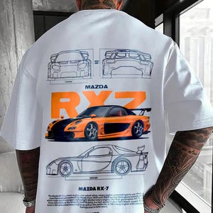 Puede incluir: Una camiseta blanca con un estampado gráfico de un coche deportivo Mazda RX-7. El diseño incluye dibujos técnicos de líneas del coche, el texto "MAZDA RX-7" en letras grandes de color naranja y un párrafo detallado con texto histórico sobre el vehículo.