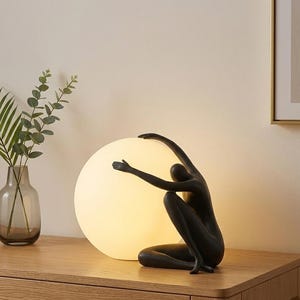 May include: A black sculpture of a person embracing a large, glowing orb, likely a lamp. The figure is in a seated position with arms outstretched. The lamp sits on a wooden surface, with a vase of greenery to the left.